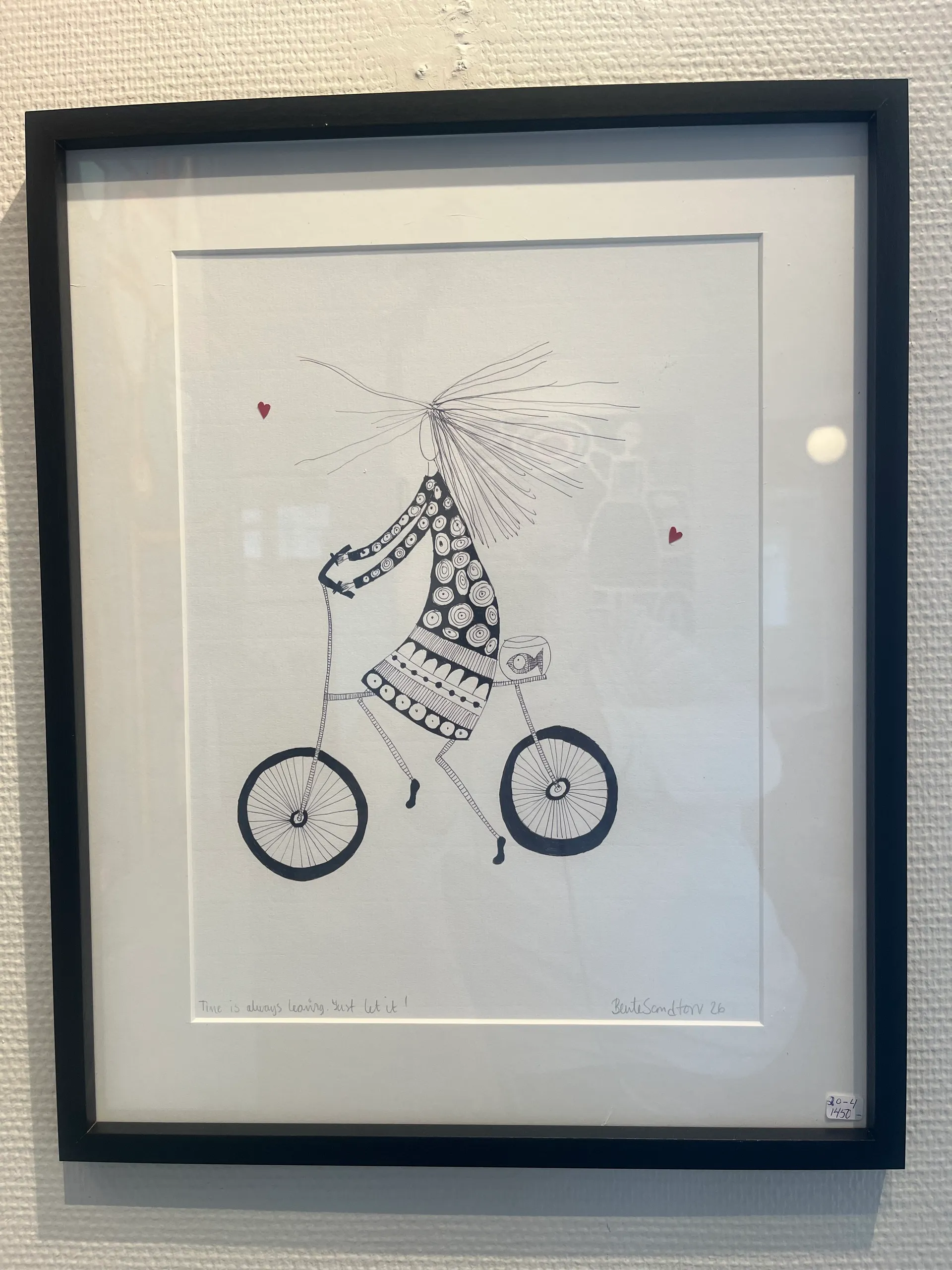 A framed black-and-white ink drawing for sale in the gallery, showing a whimsical long-haired figure in a patterned dress riding a bicycle, hand-titled 'Time is always leaving. Just let it!' and signed Bente Sandtorv