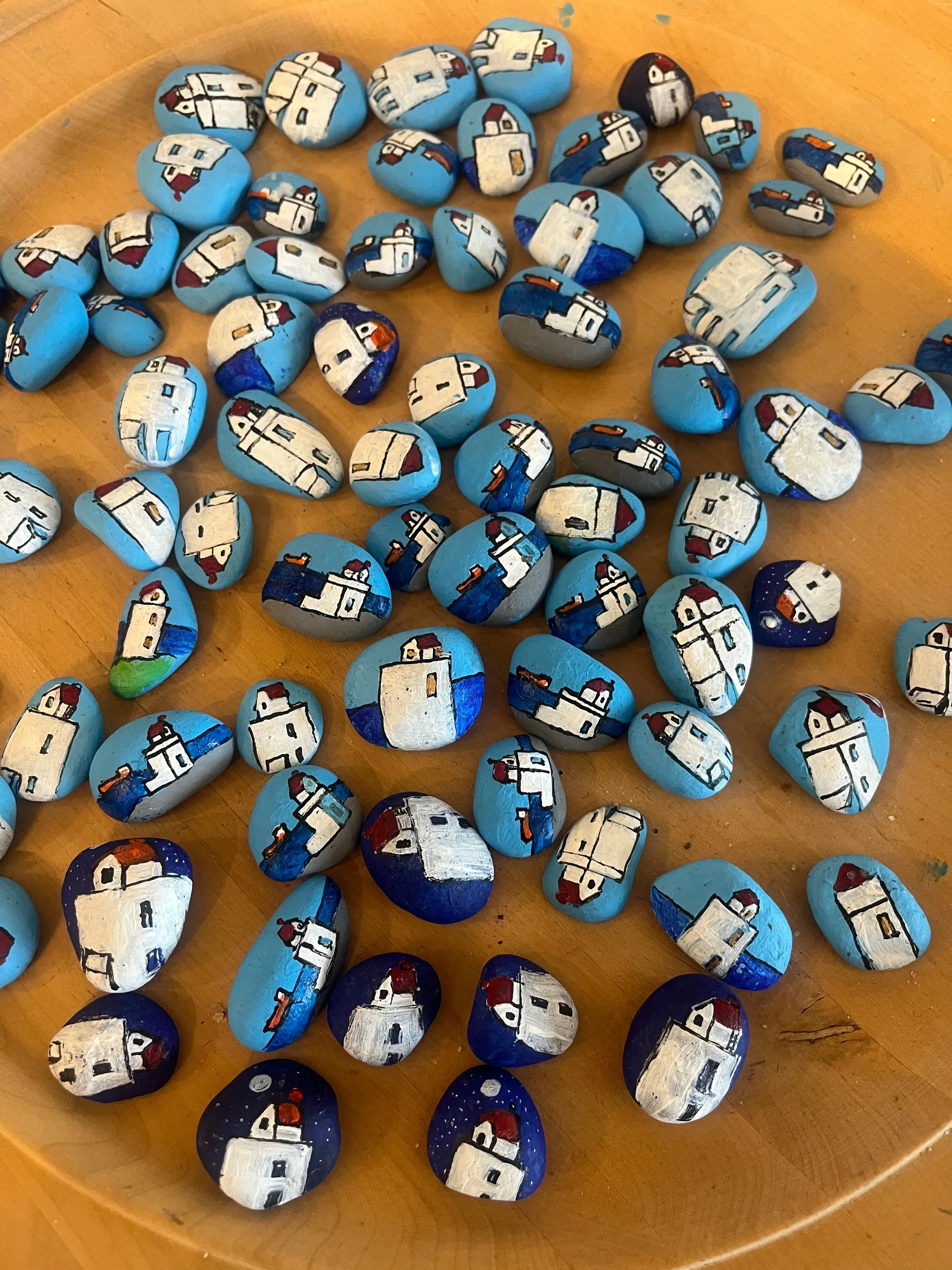 A wooden bowl filled with dozens of small beach stones, each hand-painted with white coastal houses and red roofs against blue and turquoise backgrounds
