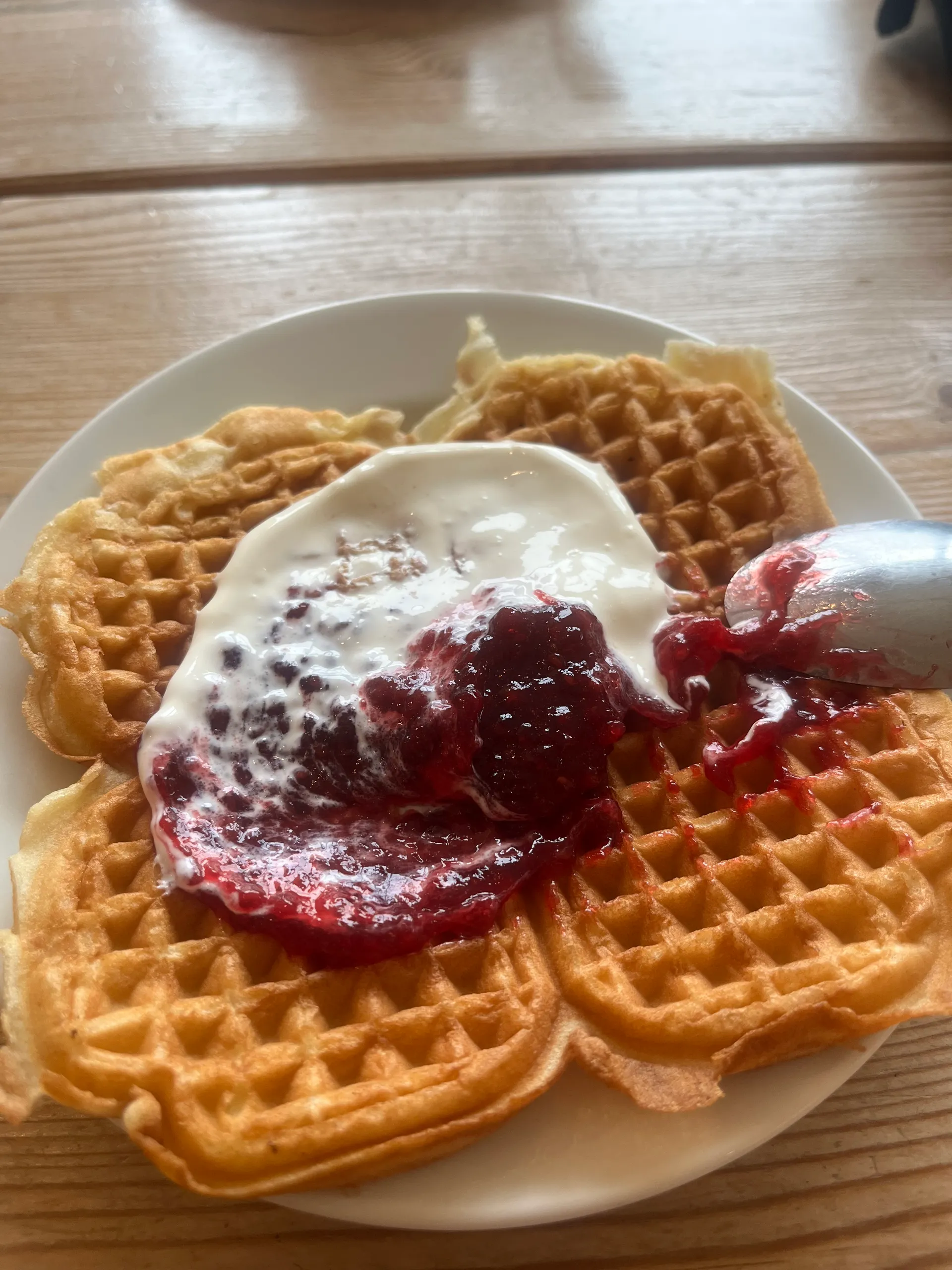 A plate of golden heart-shaped Norwegian waffles topped with a generous dollop of soured cream and red berry jam, with a jam spoon resting on the plate