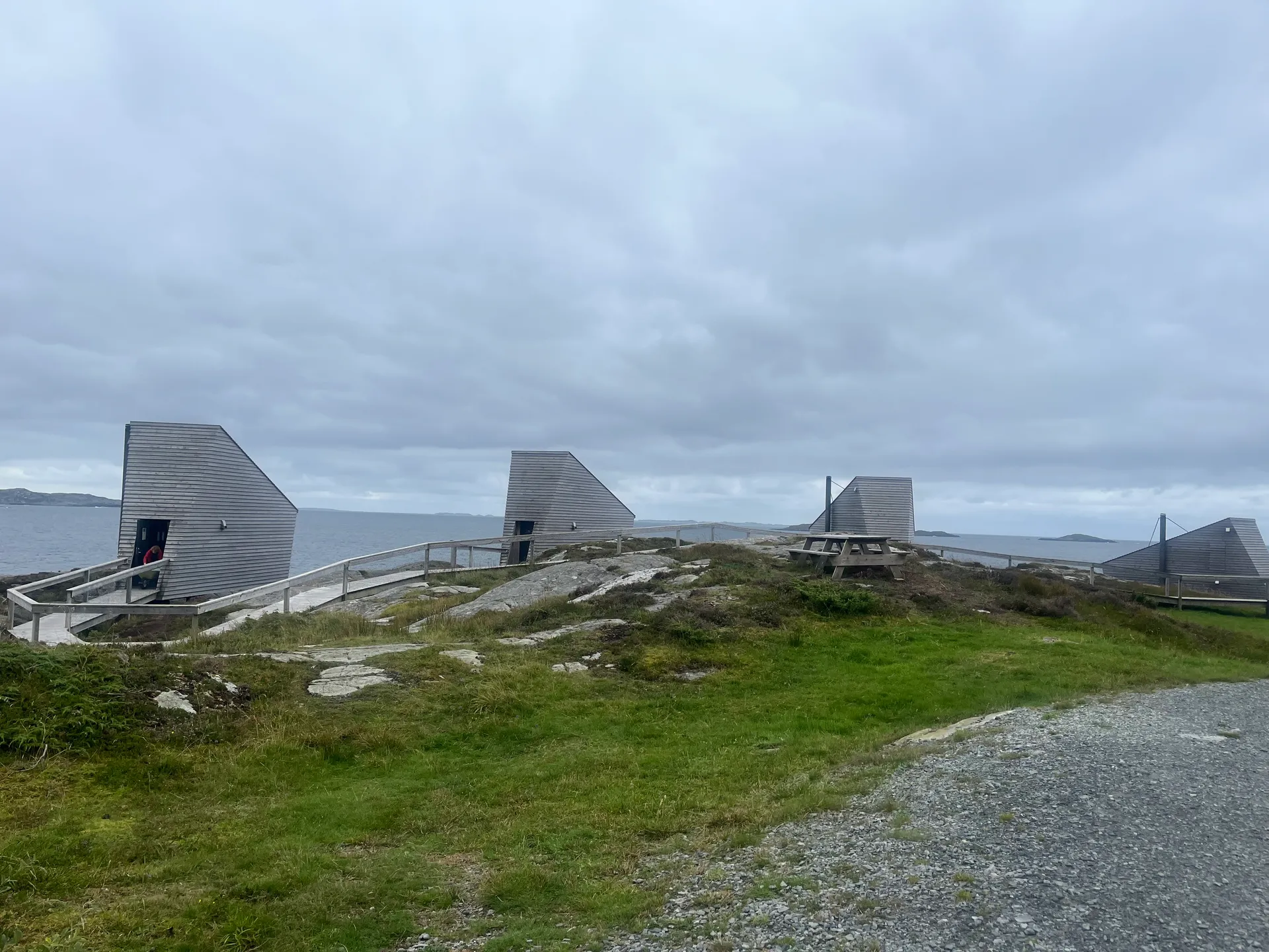 Four of the angular grey-timber Floke cottages spread across a rocky, grassy headland at Ryvarden, linked by a wooden boardwalk above the sea under a moody overcast sky