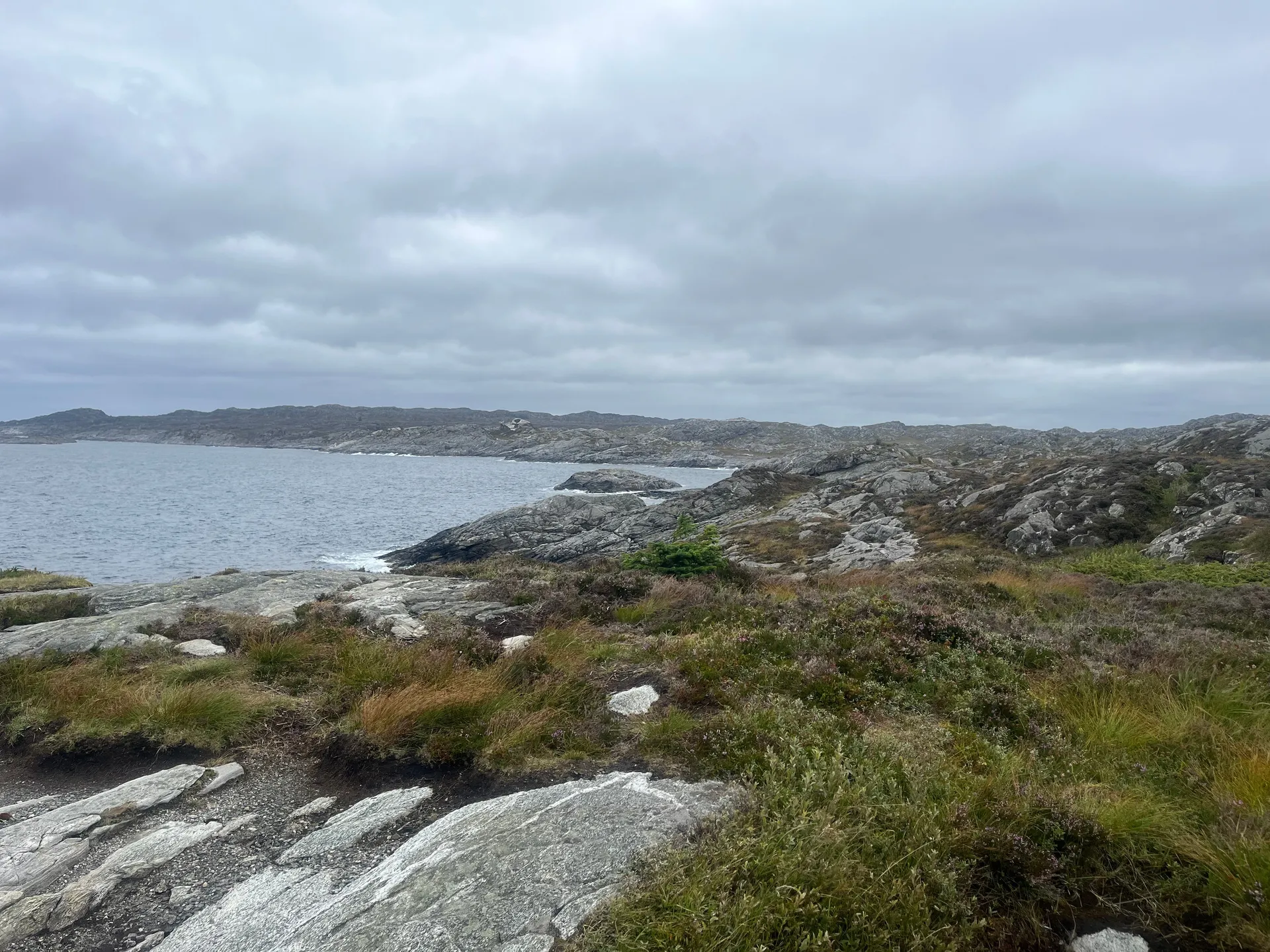 A sweeping view along the Ryvarden peninsula — granite outcrops, grass and heather in the foreground with the grey sea breaking against rocks and a low island ridge on the horizon under an overcast sky