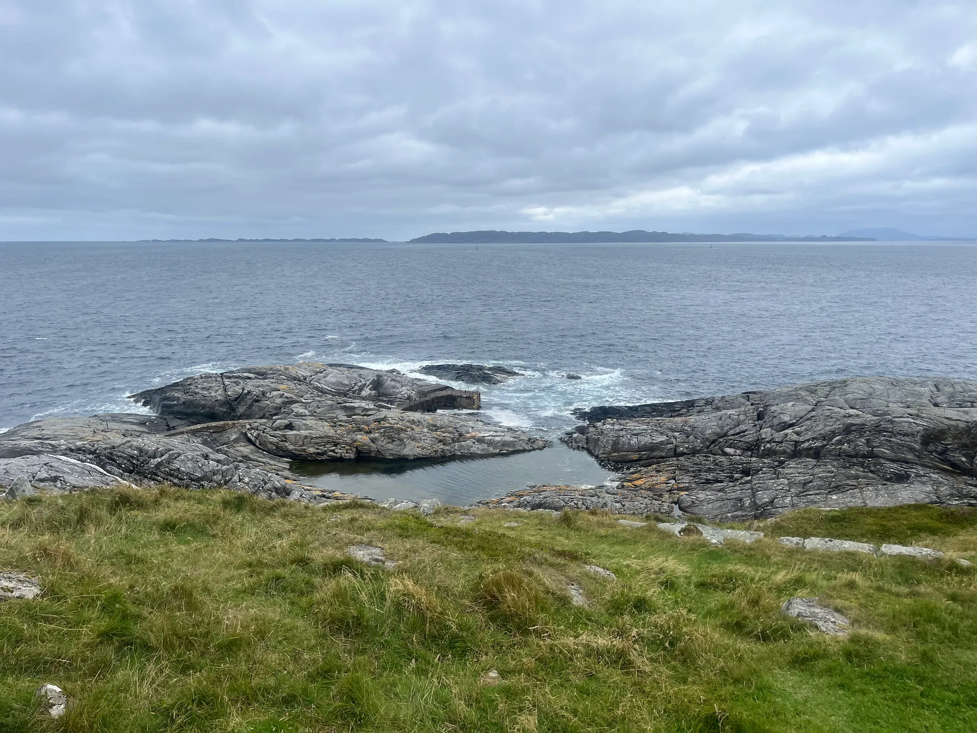 Looking out to sea from a grassy headland at Ryvarden, over layered grey rock formations and a calm tidal pool, with a distant island on the horizon under a cloudy sky