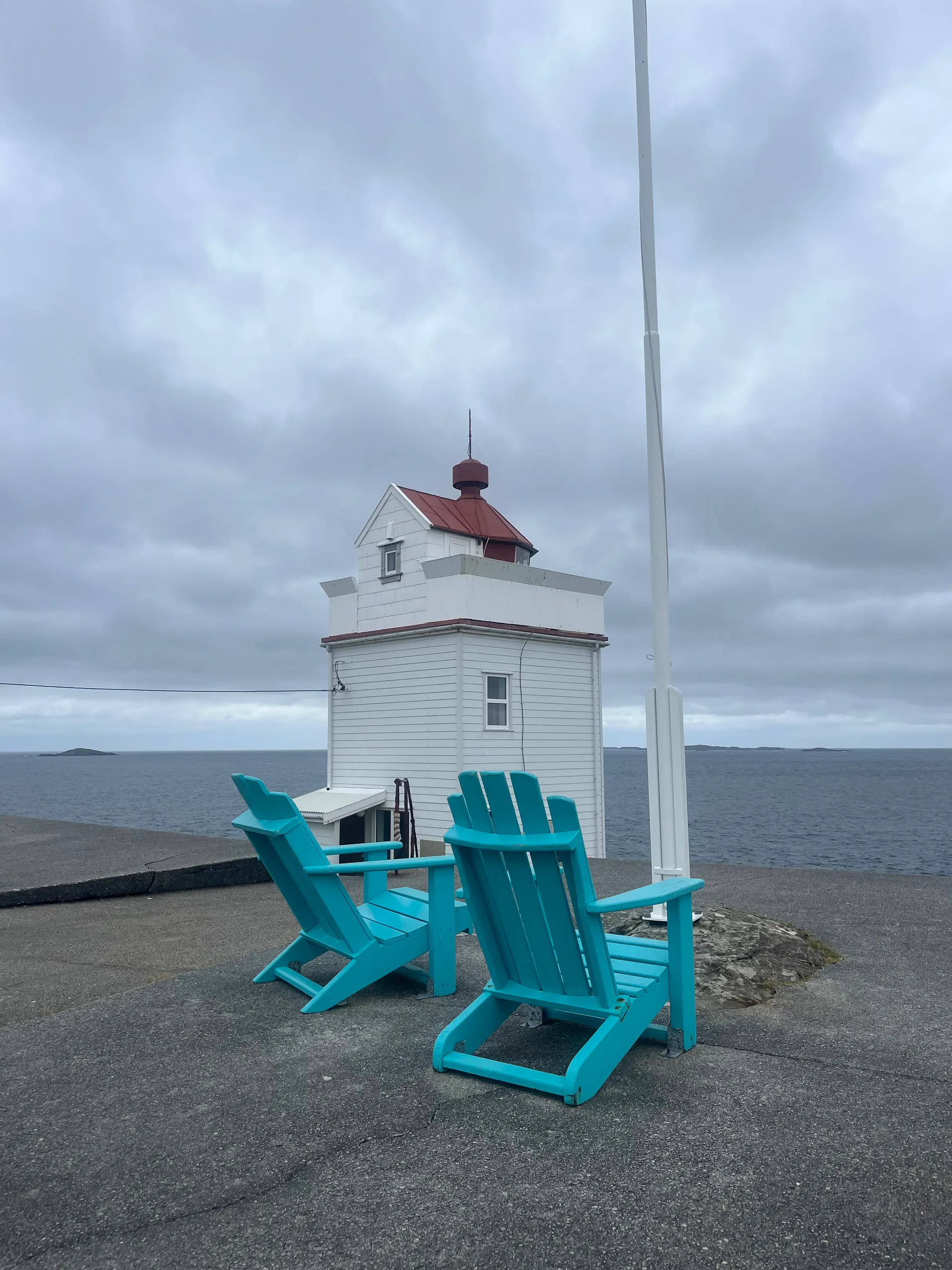 Two turquoise Adirondack deck chairs on the concrete platform at Ryvarden, framing the white lighthouse tower with its red lantern roof against a moody grey sky and the open sea
