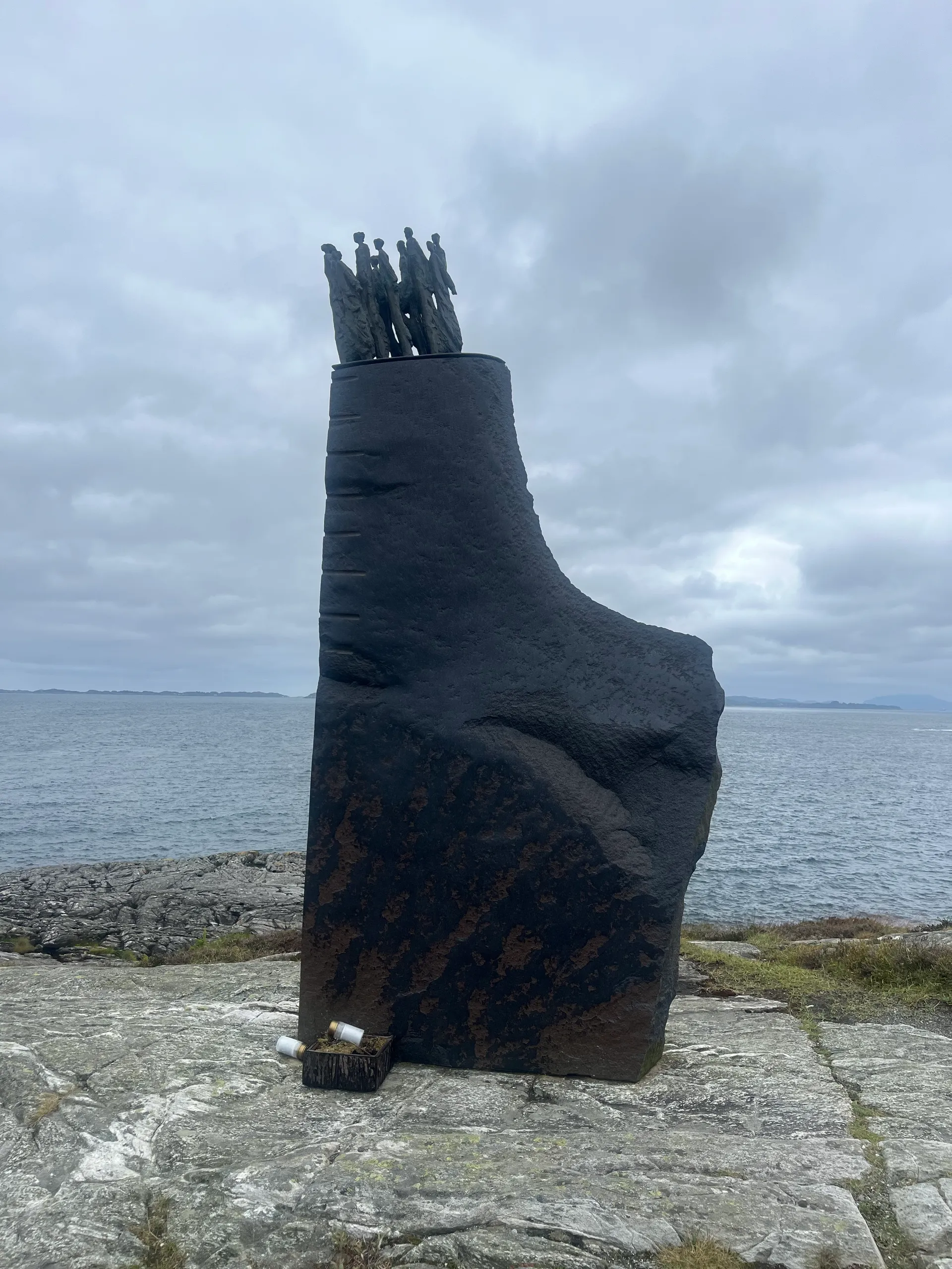 The Sleipner memorial at Ryvarden — a tall, dark rough-hewn monument shaped like a cliff, topped with a cluster of small bronze human figures, standing on the rocky coast overlooking the grey sea under an overcast sky