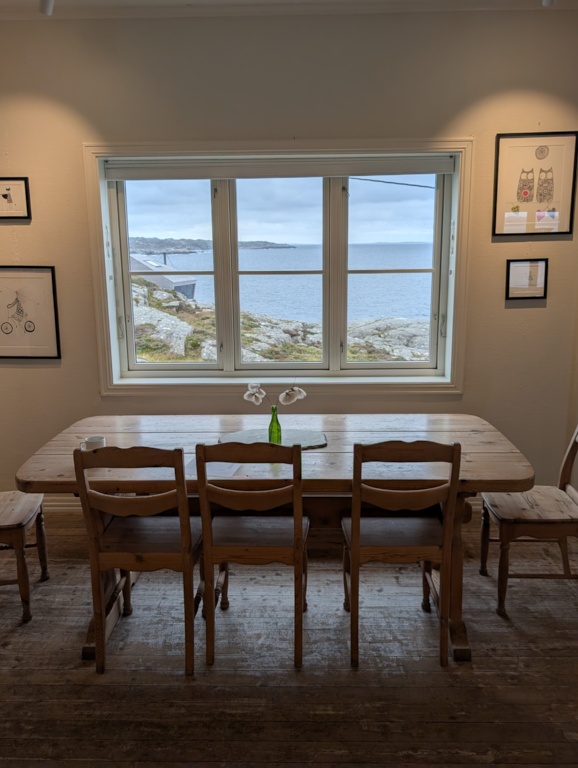 A rustic wooden dining table and chairs in the Ryvarden cafeteria beneath a large window framing the open sea and rocky coastline