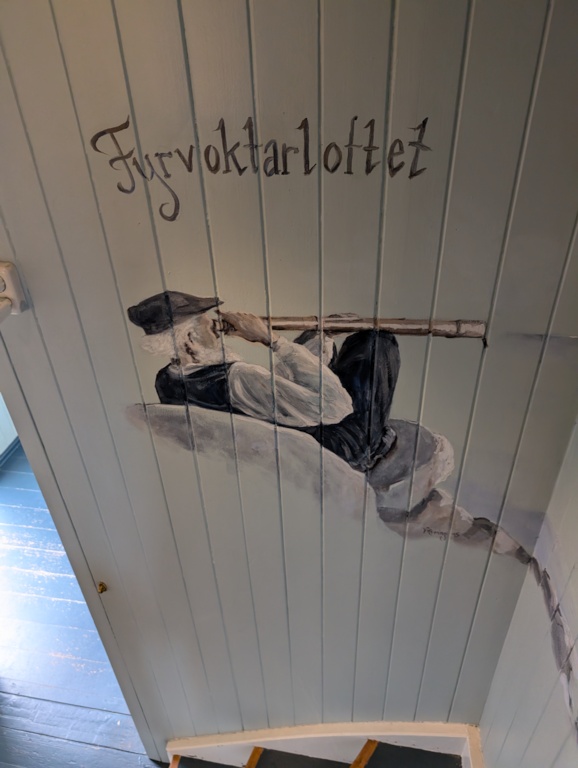 A hand-painted mural on the pale wooden plank ceiling of the staircase up to the keeper's quarters, showing a fisherman in dark clothes and a cap reclining against rocks with a long pole, beneath the painted word 'Fyrvoktarloftet'