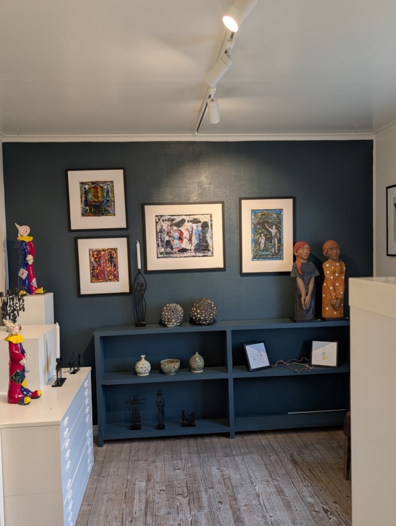 An intimate gallery corner at Ryvarden with a deep teal wall, framed colorful prints, painted ceramic figures and carved wooden sculptures on display shelves