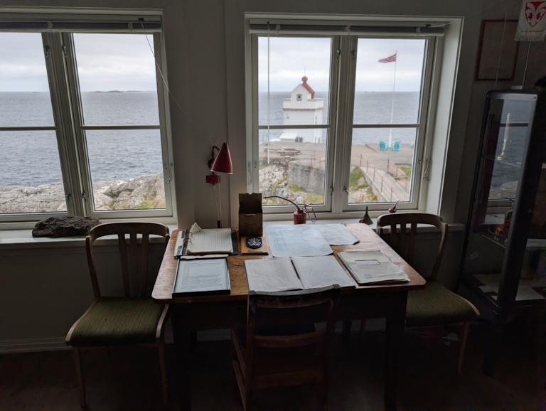The lighthouse keeper's desk at Ryvarden, spread with old logbooks and papers beneath tall windows looking out to the white lighthouse tower and a flying Norwegian flag