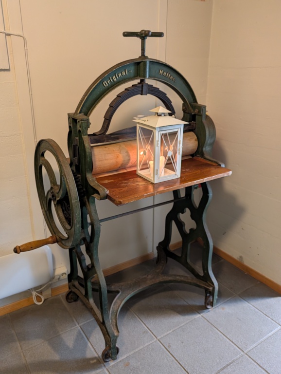 An antique green cast-iron laundry mangle with a wooden roller and spoked flywheel, on display in the keeper's building with a white lantern set on top